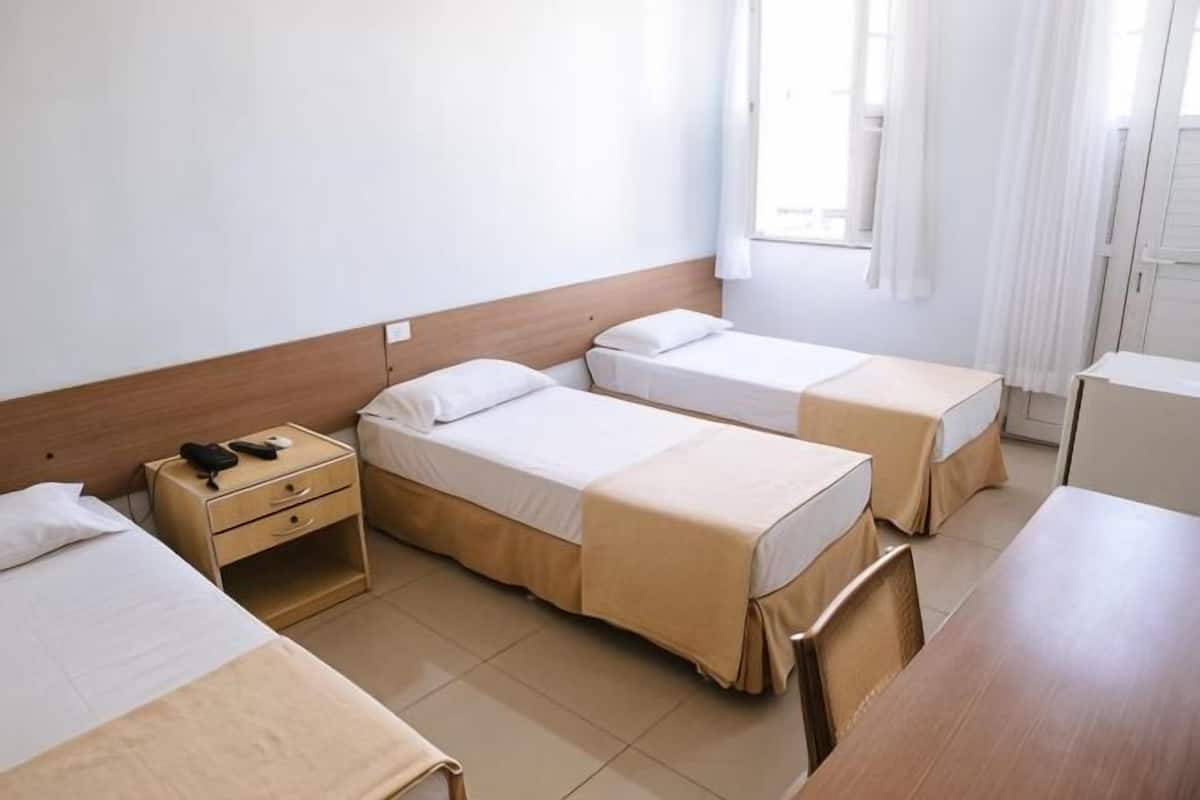 A hotel room with three single beds, a wooden headboard, a bedside table with a phone, and a window with curtains.
