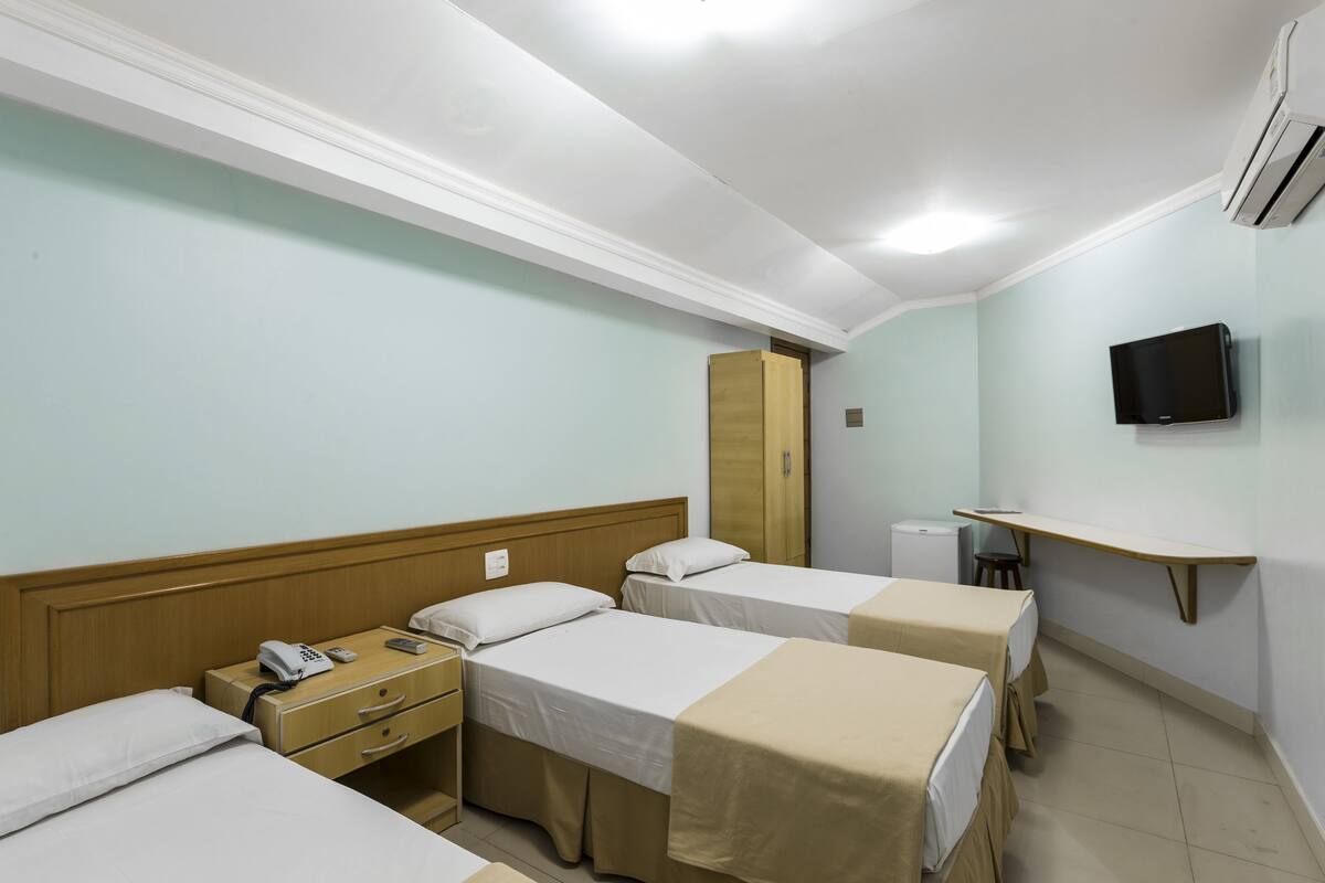 A hotel room with three single beds, a wooden wardrobe, a desk with a chair, a television mounted on the wall, and an air conditioning unit.