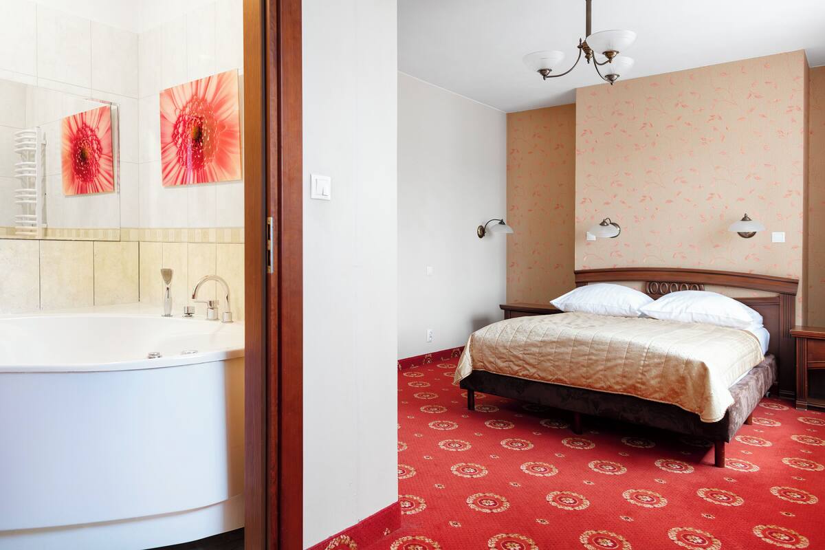 A hotel room with a large bathtub, a bed with a red patterned carpet, and a wall with floral wallpaper.