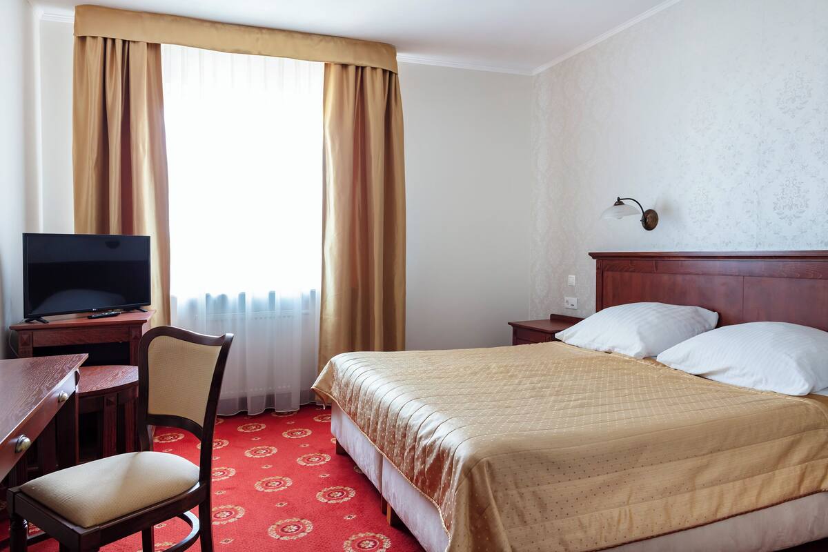 A hotel room with a bed, a desk with a television, a chair, and a nightstand with a lamp.
