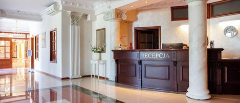Zona de recepción con suelo de mármol, un mostrador de recepción con la etiqueta “RECEPCIA” y un espejo grande colgando sobre él.