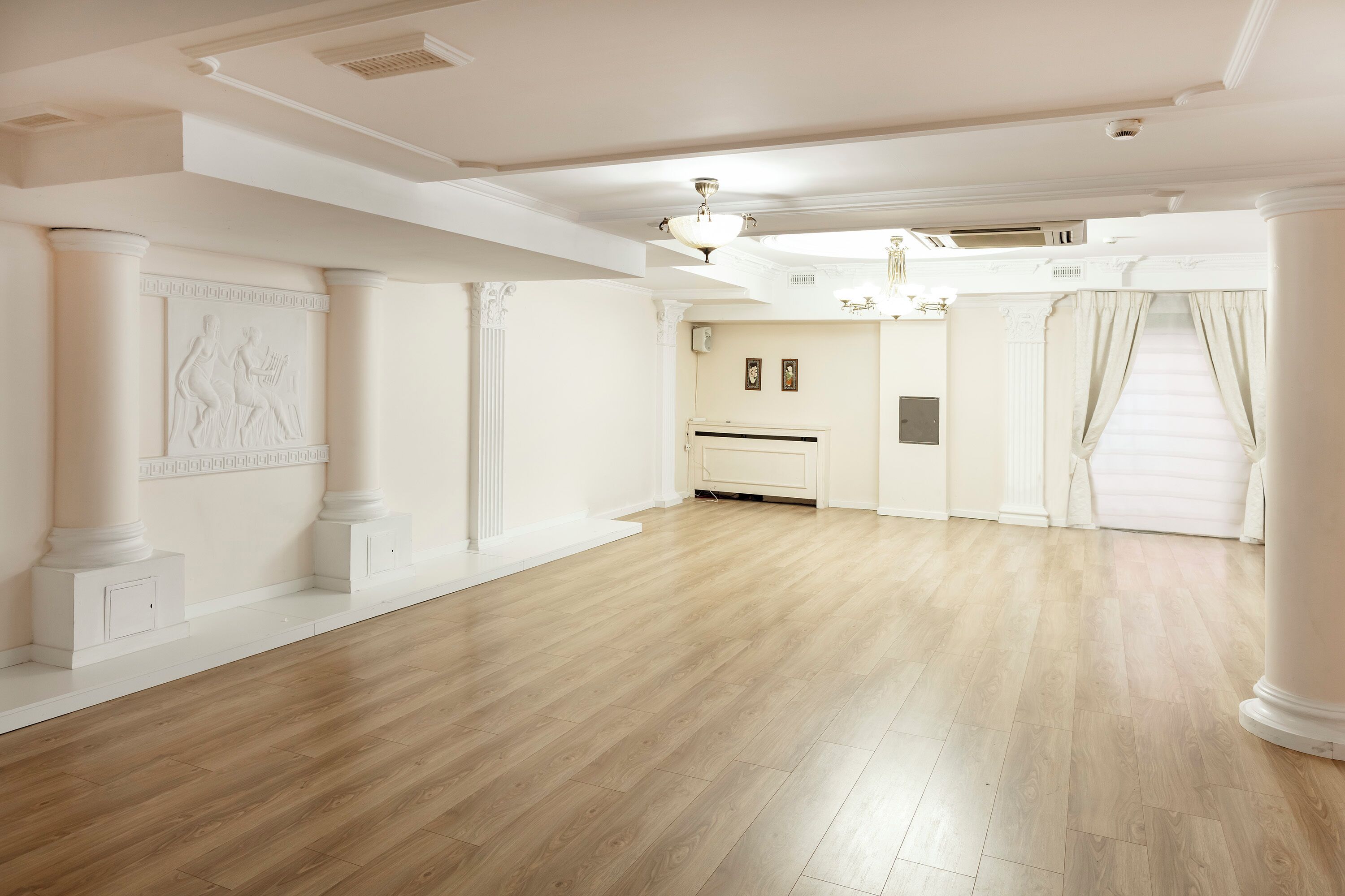 A spacious room with a wooden floor, white walls, and columns. There is a chandelier, a fireplace, and a large window with curtains.