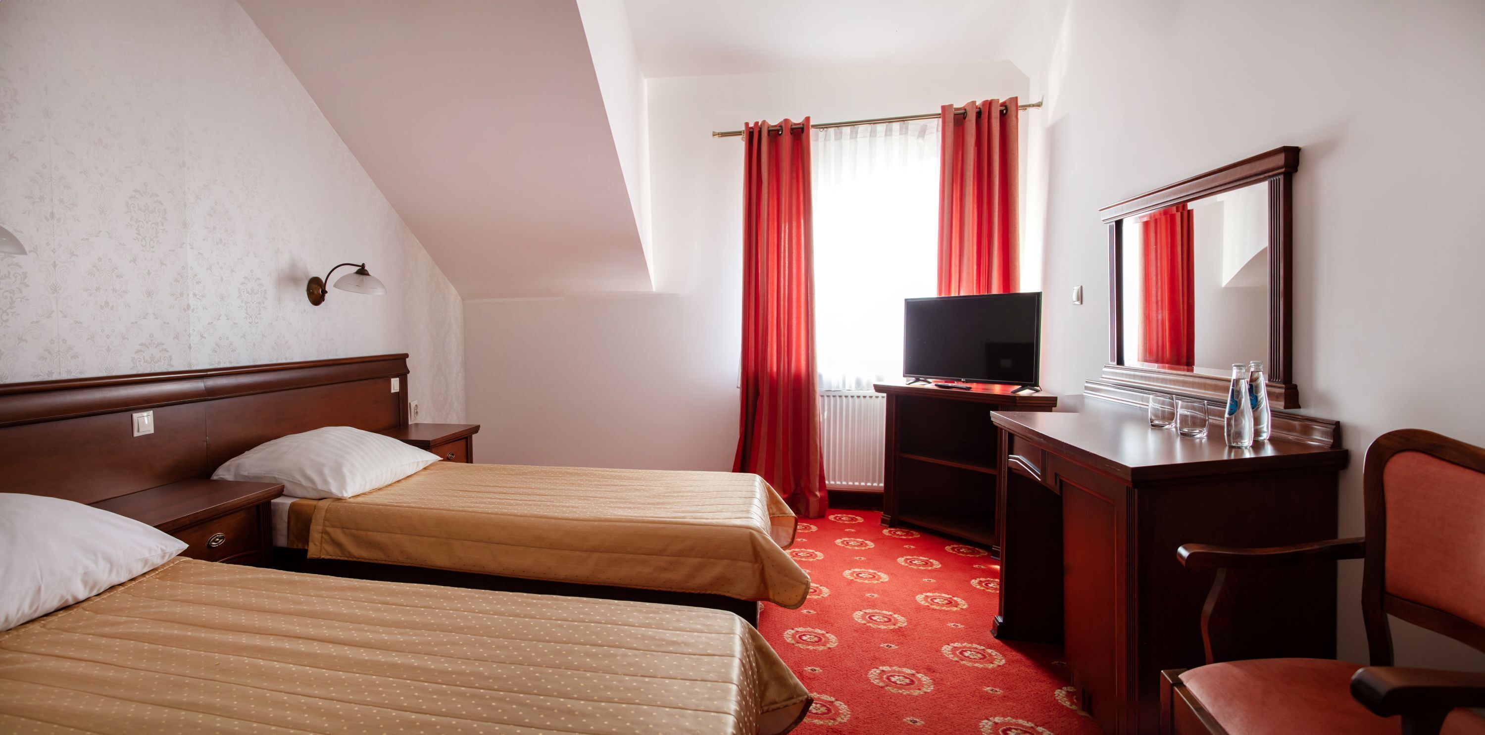 A hotel room with two beds, a desk with a TV, a mirror, and a red carpet.