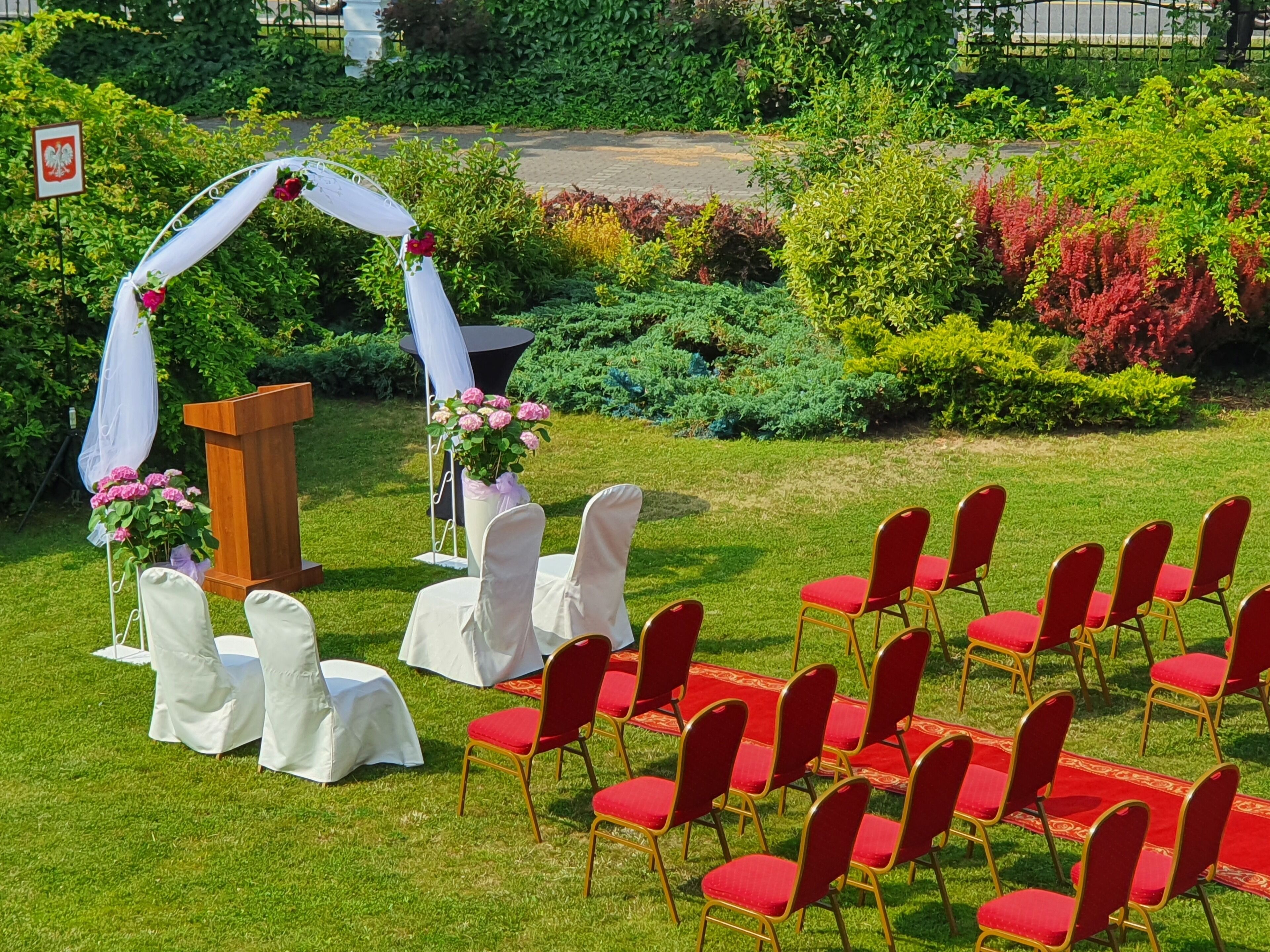 Une cérémonie de mariage en plein air, avec un podium en bois, des chaises blanches et un tapis rouge.