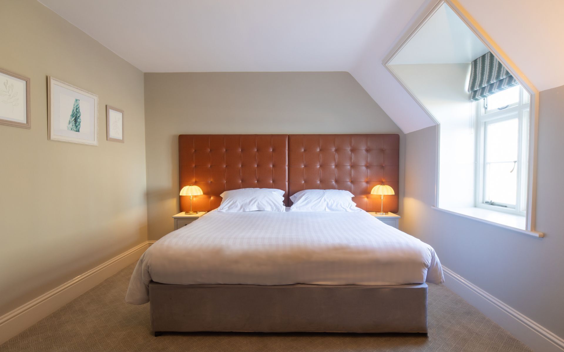 A neatly made bed with a tufted headboard, two bedside lamps, and a large window with a view of the sky.