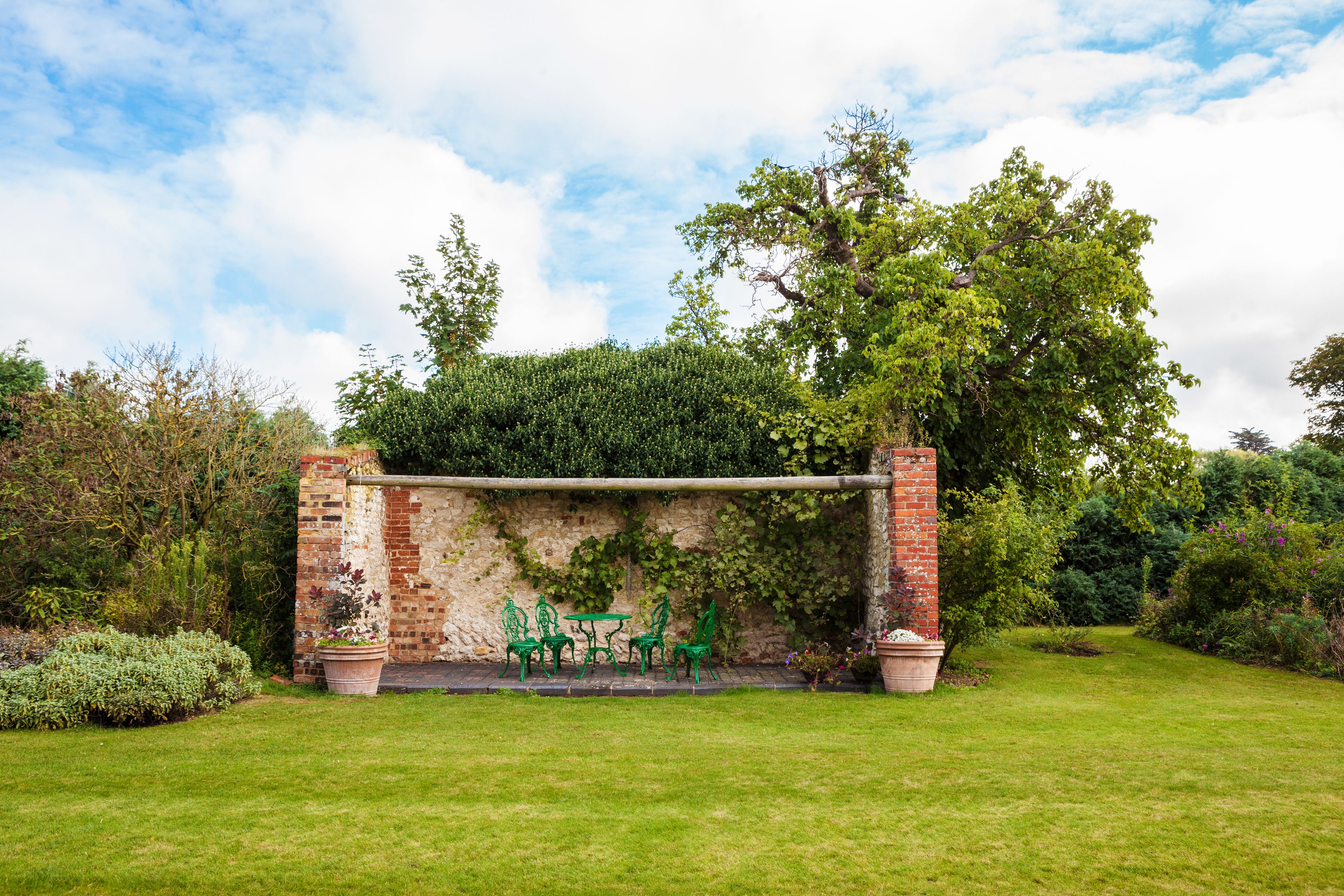 A brick pergola with green metal chairs and tables in a garden setting.