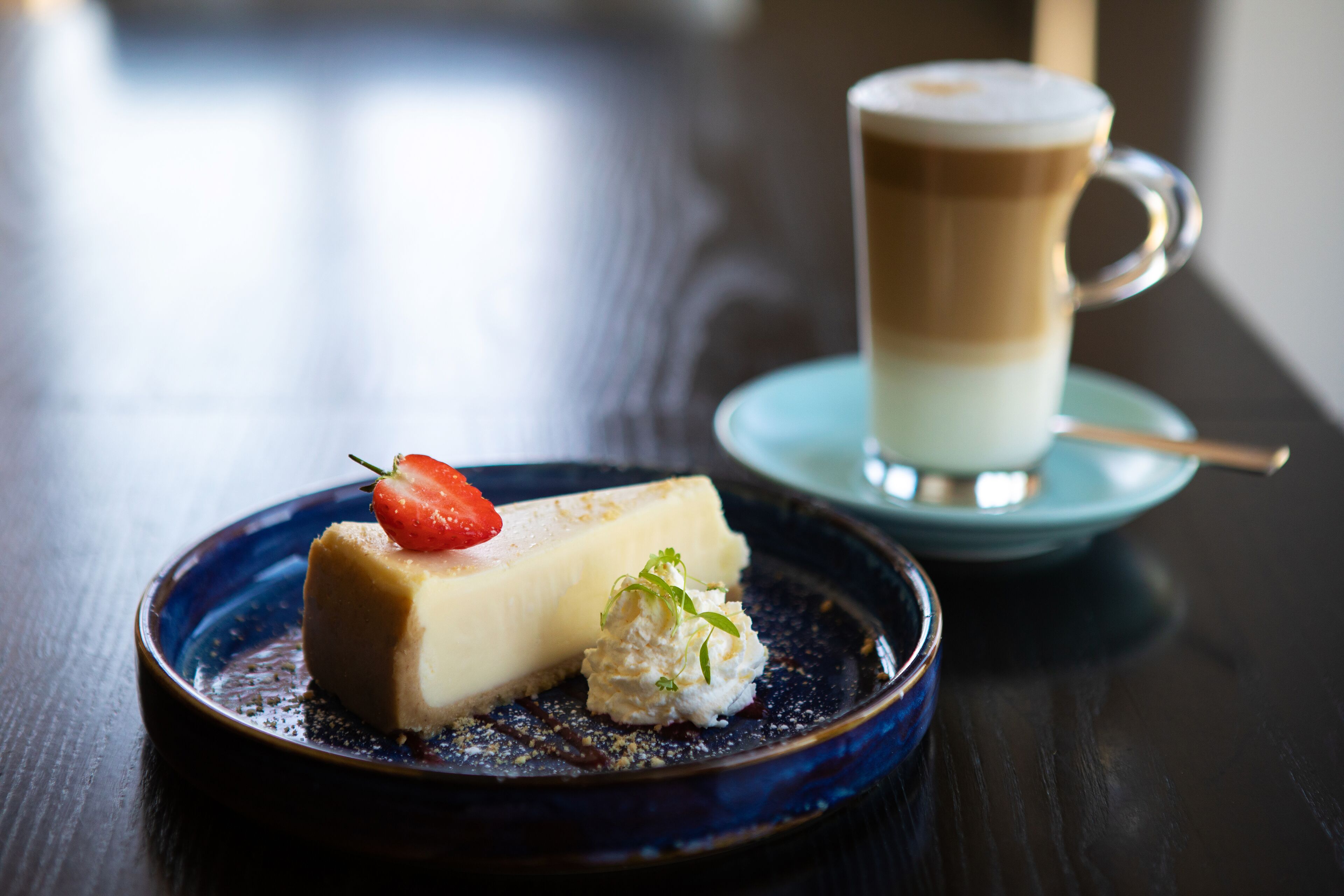 A dessert plate with a slice of cheesecake and a strawberry on a dark blue plate, accompanied by a cup of coffee on a saucer.