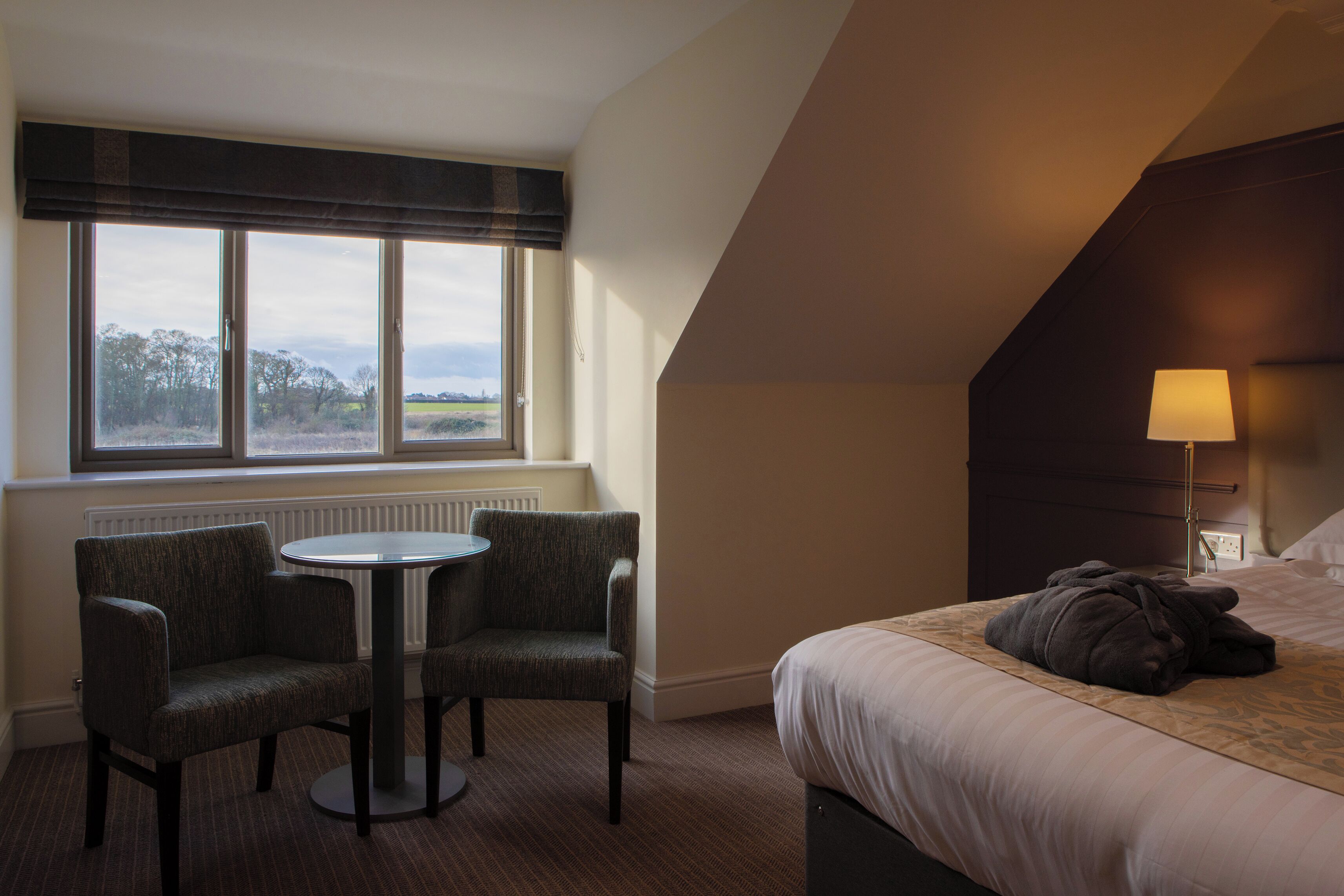 Ein Hotelzimmer mit Bett, Tisch mit zwei Stühlen, Fenster mit Blick ins Grüne und einer Lampe auf dem Nachttisch.