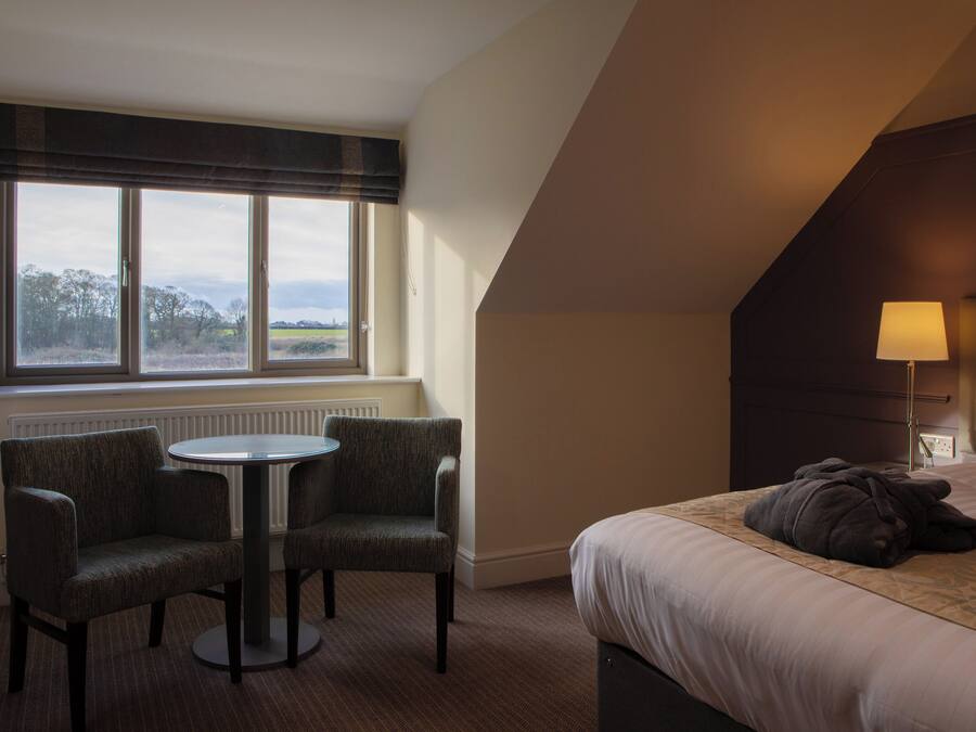 Ein Hotelzimmer mit Bett, Tisch mit zwei Stühlen, Fenster mit Blick ins Grüne und einer Lampe auf dem Nachttisch.