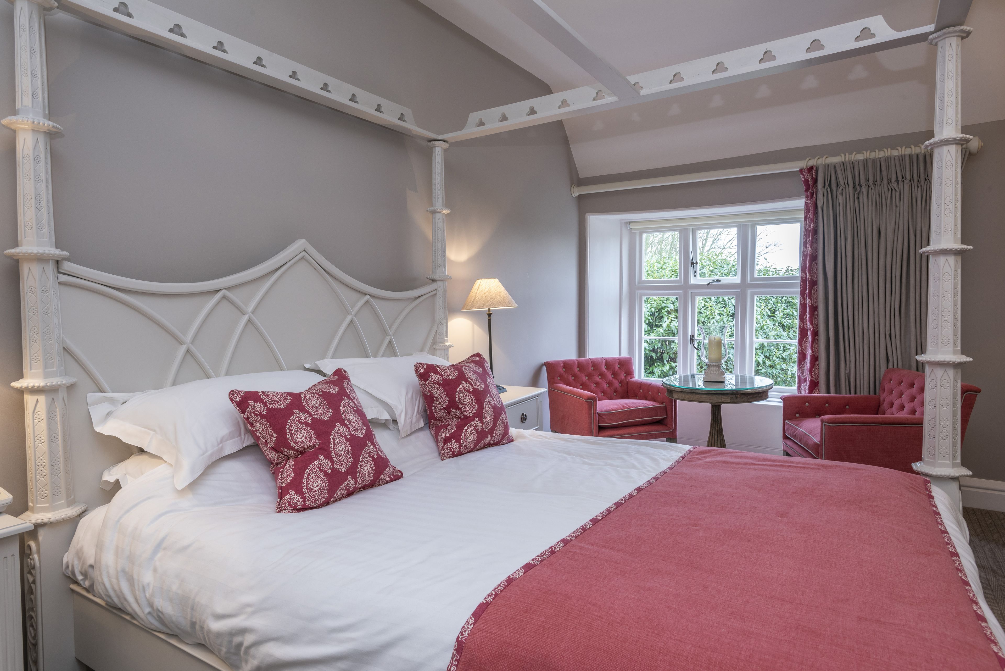 A four-poster bed with white and red bedding, a small round table with a lamp, and two red chairs in a room with a large window and a view of greenery.