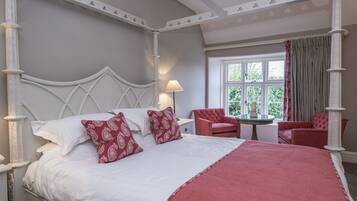 A four-poster bed with white and red bedding, a small round table with a lamp, and two red chairs in a room with a large window and a view of greenery.