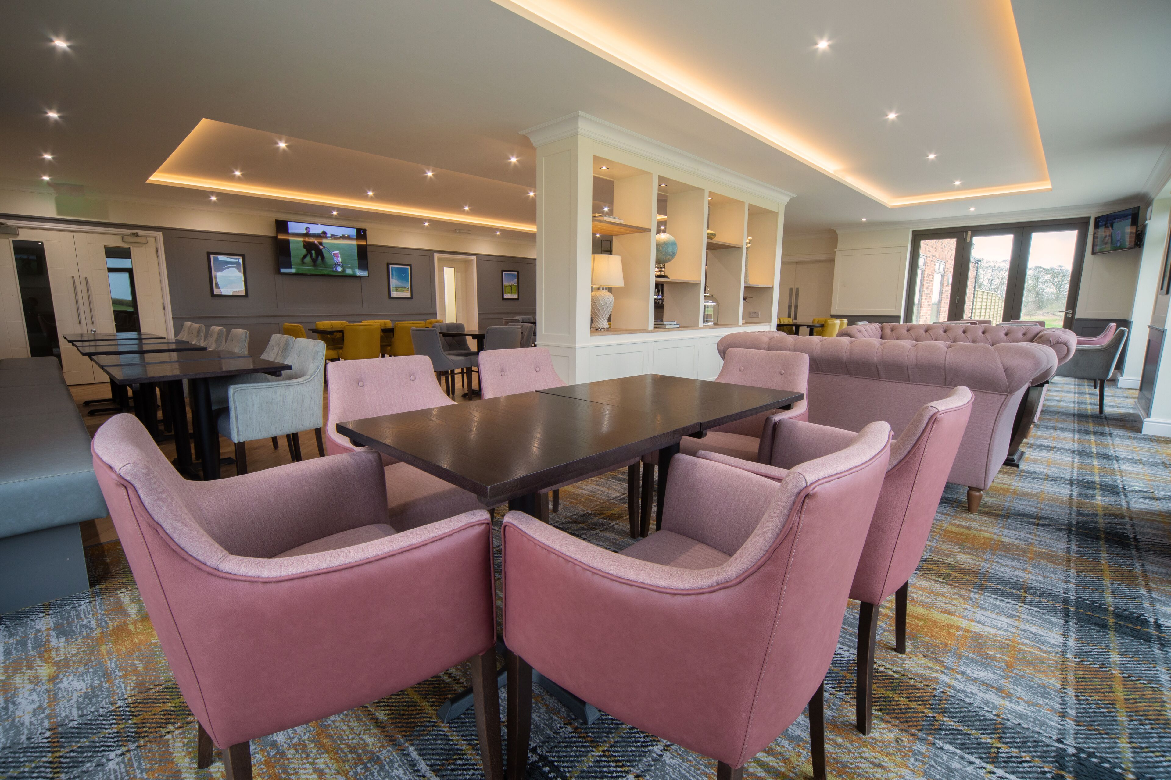 A modern hotel lobby with a large dining table, comfortable seating, a television, and a view of the outdoors.