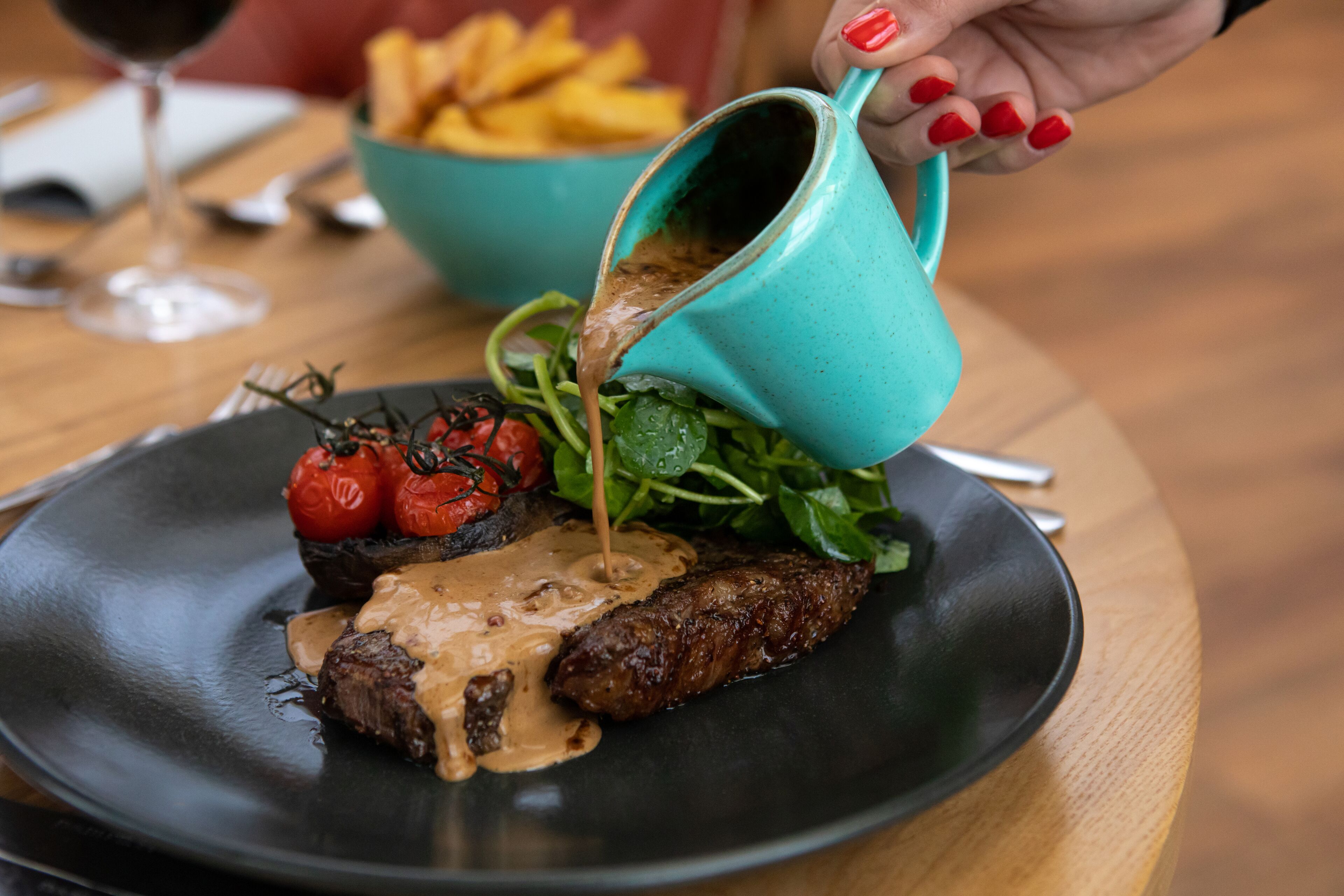 A plate of food with a steak, vegetables, and sauce, being poured from a turquoise pitcher.