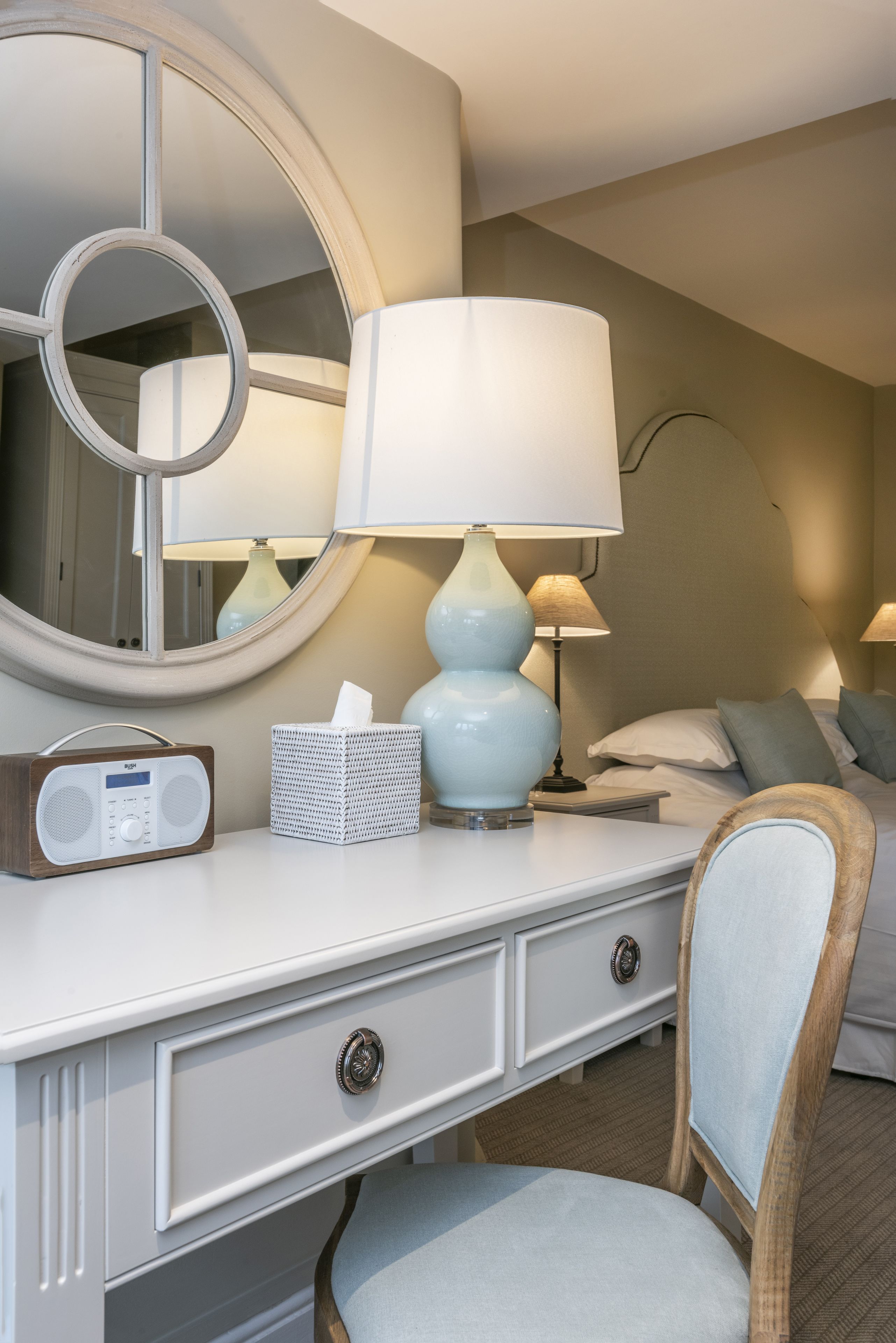 A neatly arranged hotel room with a white dresser, a round mirror, a lamp, a radio, and a chair.