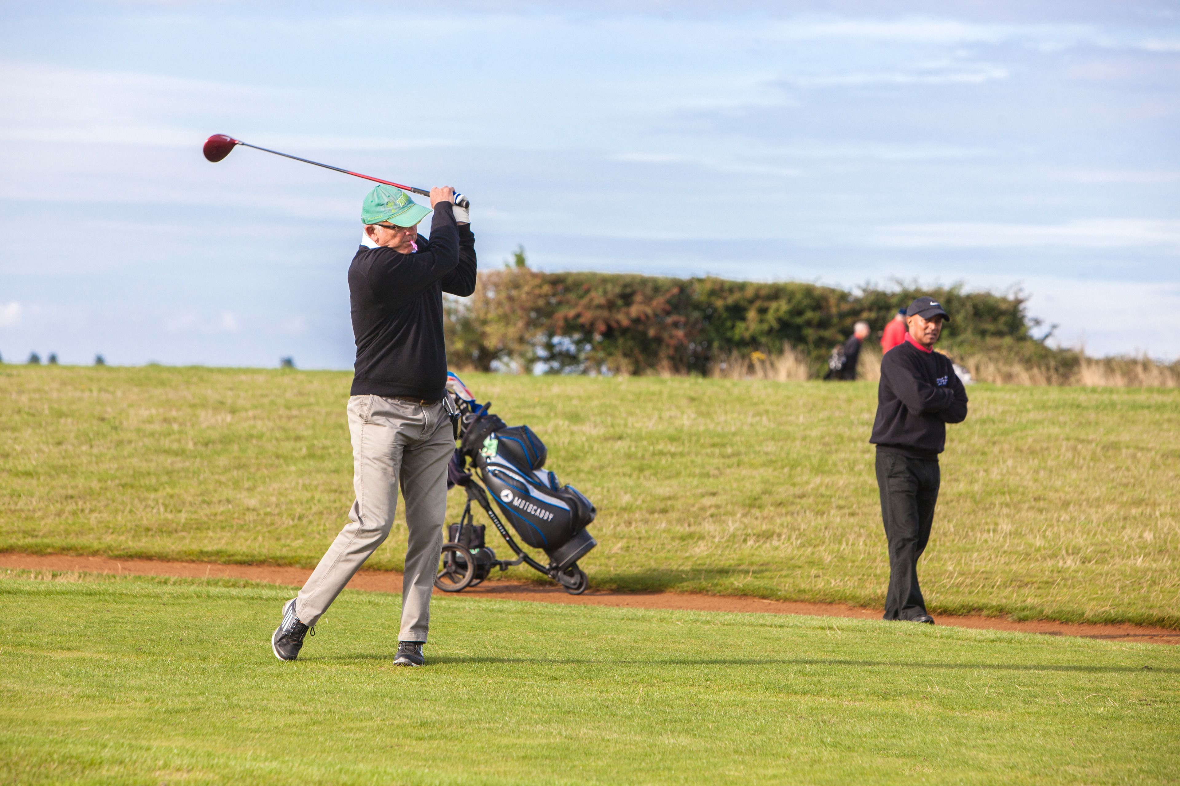 A golf course with a player in mid-swing and another person observing.