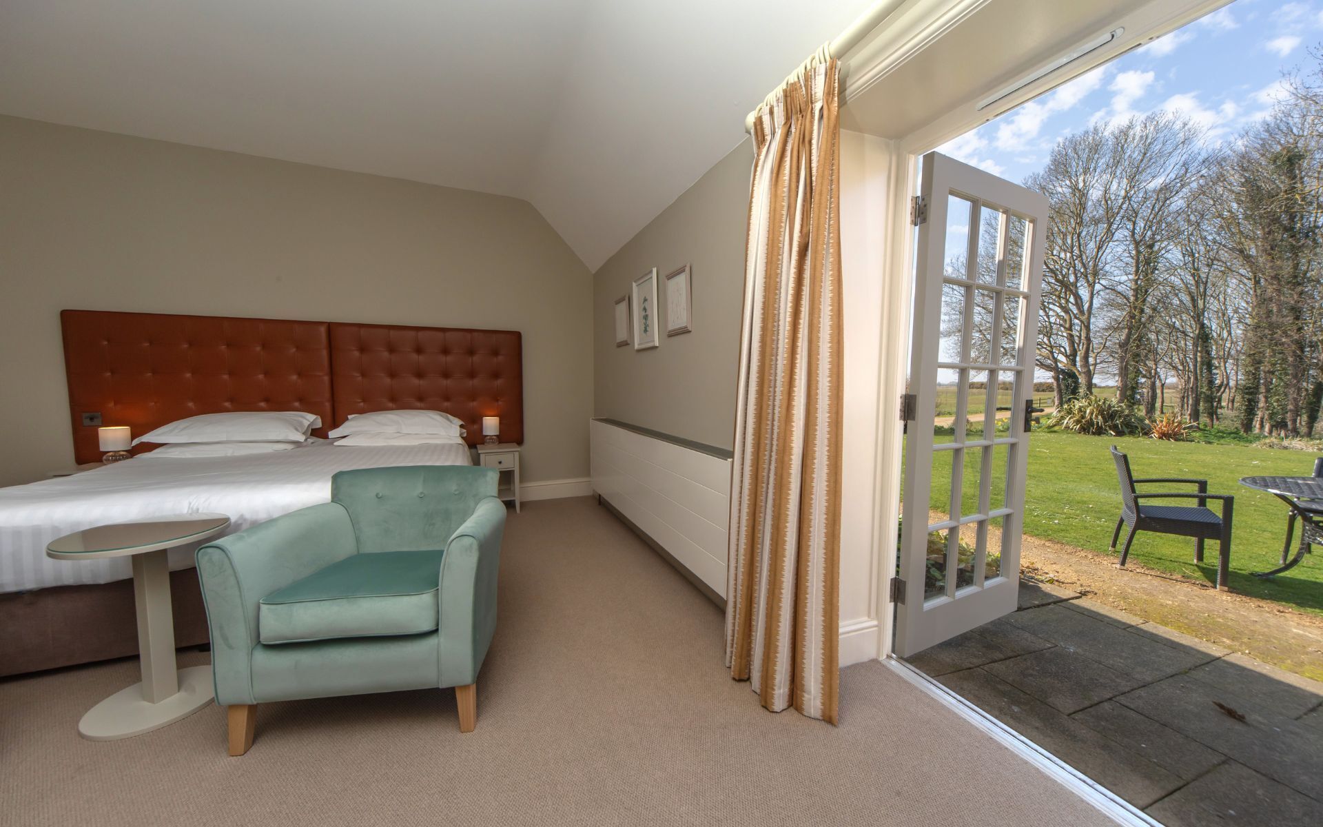 A hotel room with a bed, a small table, a chair, and a view of the garden through a glass door.