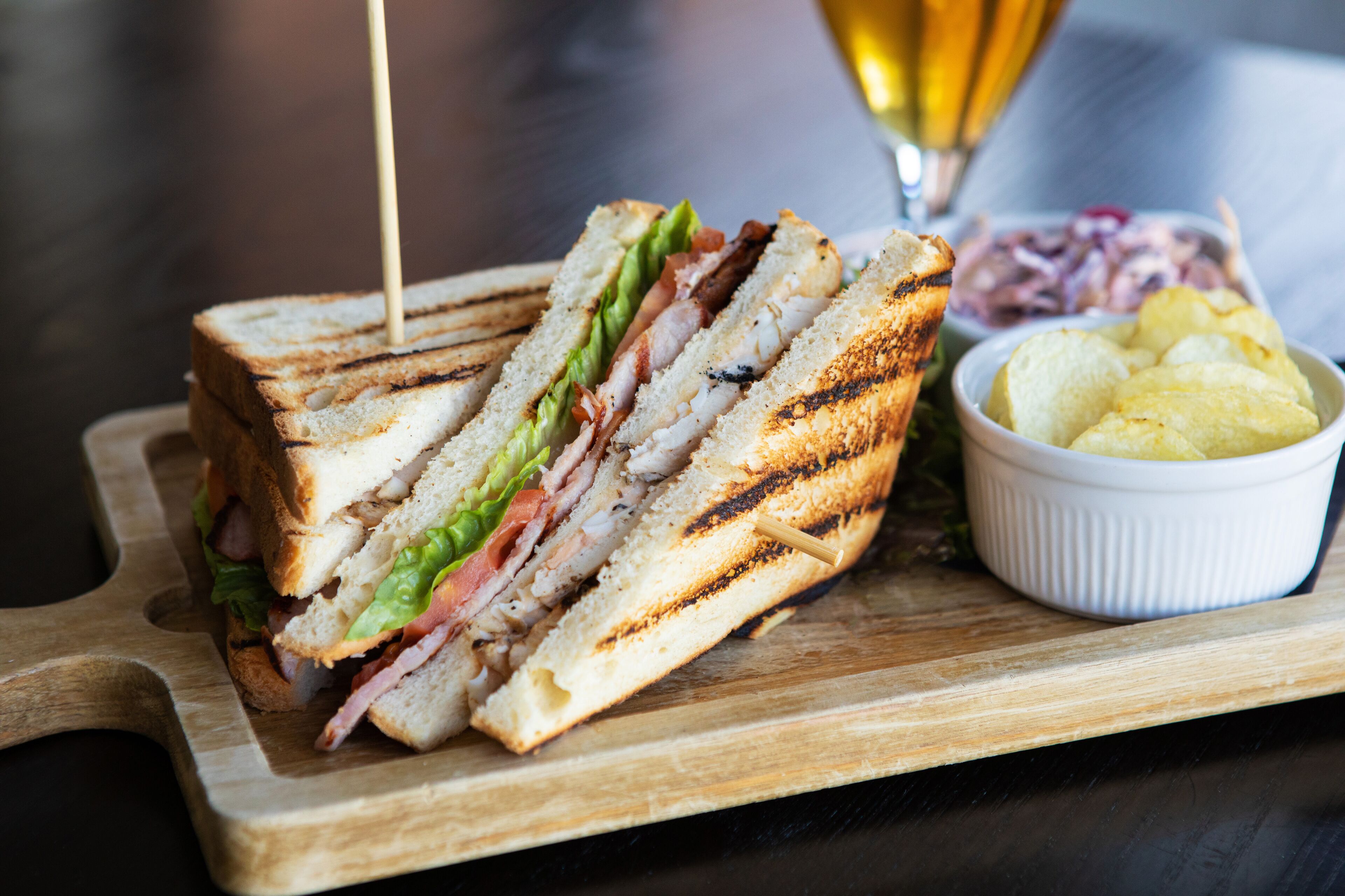 A sandwich on a wooden board with a side of chips and a glass of beer.