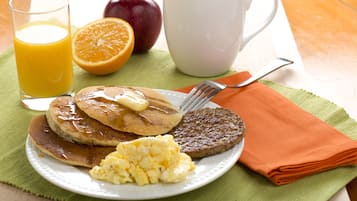 Un plato de desayuno con tortitas, salchicha, huevos revueltos y un vaso de zumo de naranja.