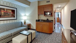 A compact hotel room with a sofa, a small coffee table, a kitchenette with wooden cabinets, a microwave, and a flat-screen TV.