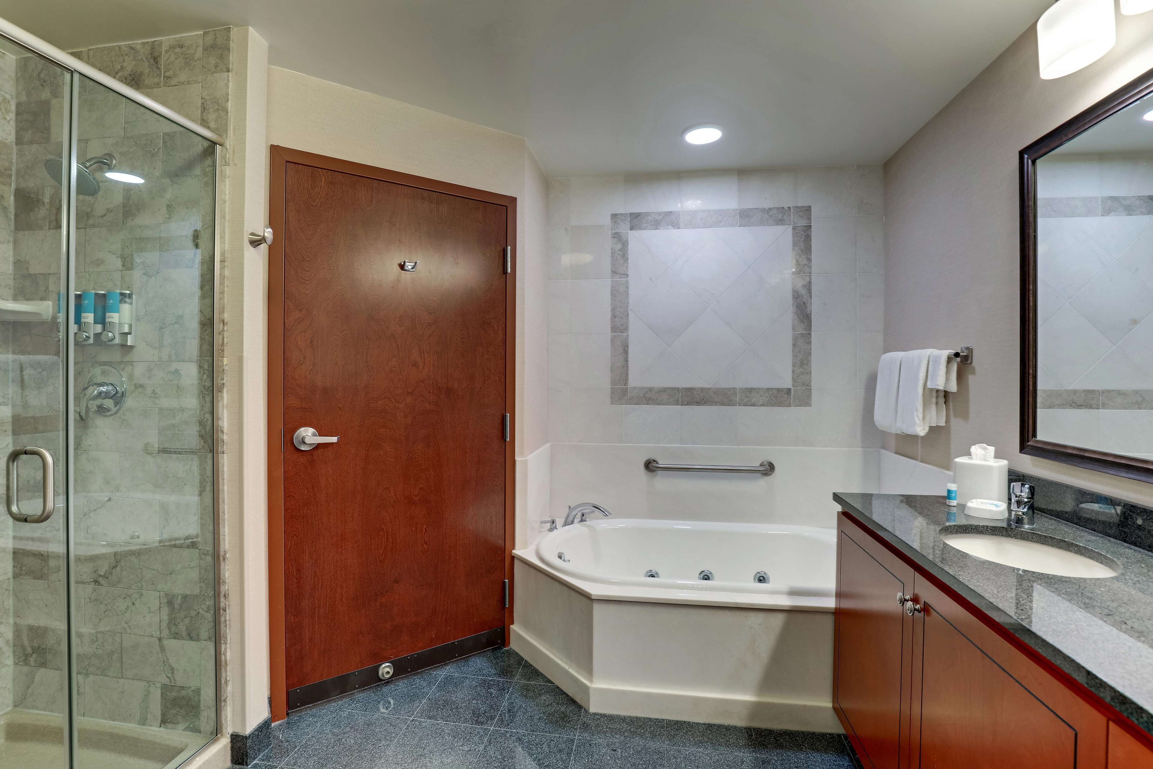 A bathroom with a glass shower enclosure, a wooden door, a bathtub, a vanity with a sink, and a mirror.