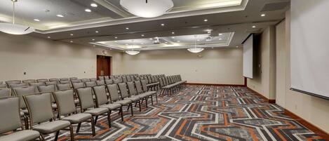 Un salón de conferencias con filas de sillas, una alfombra estampada, luces empotradas en el techo y una pantalla para proyector.