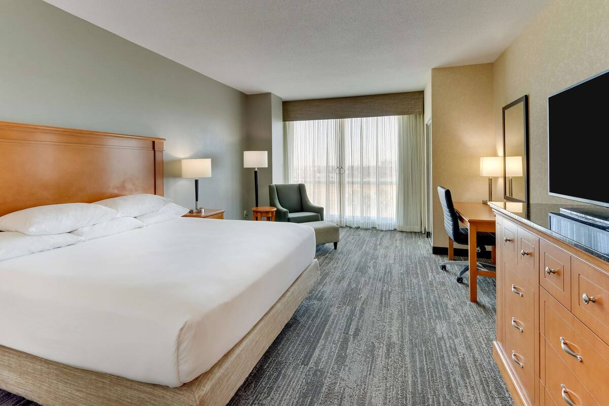 A hotel room with a large bed, a desk, a chair, a television, and a view of the city through the window.