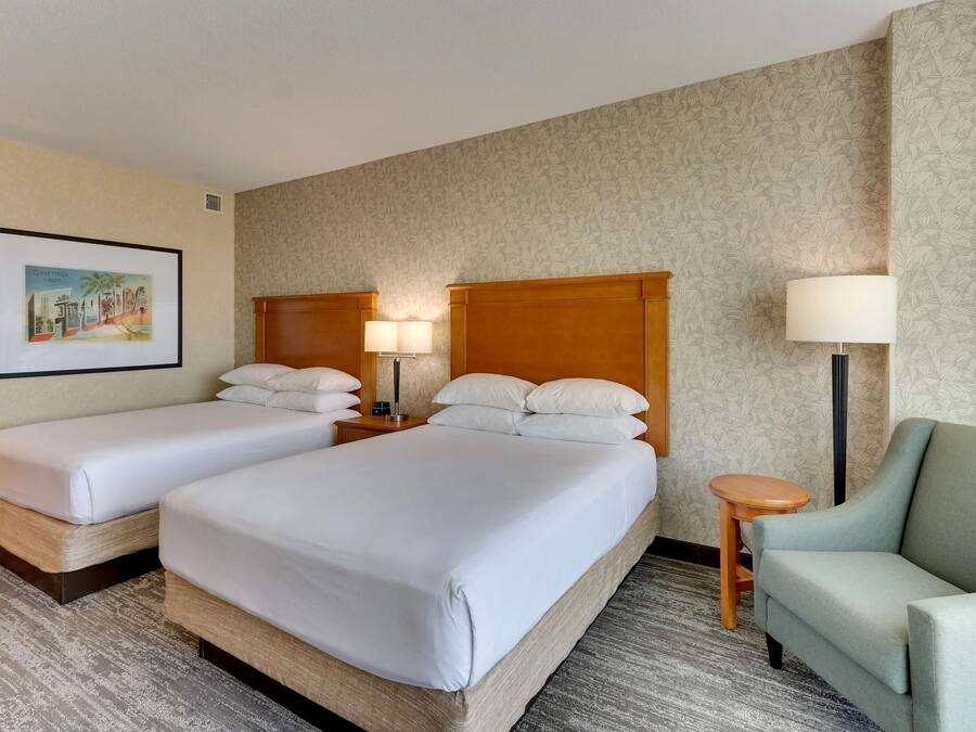 A hotel room with two single beds, a wooden headboard, a small round table, a lamp, and a chair.