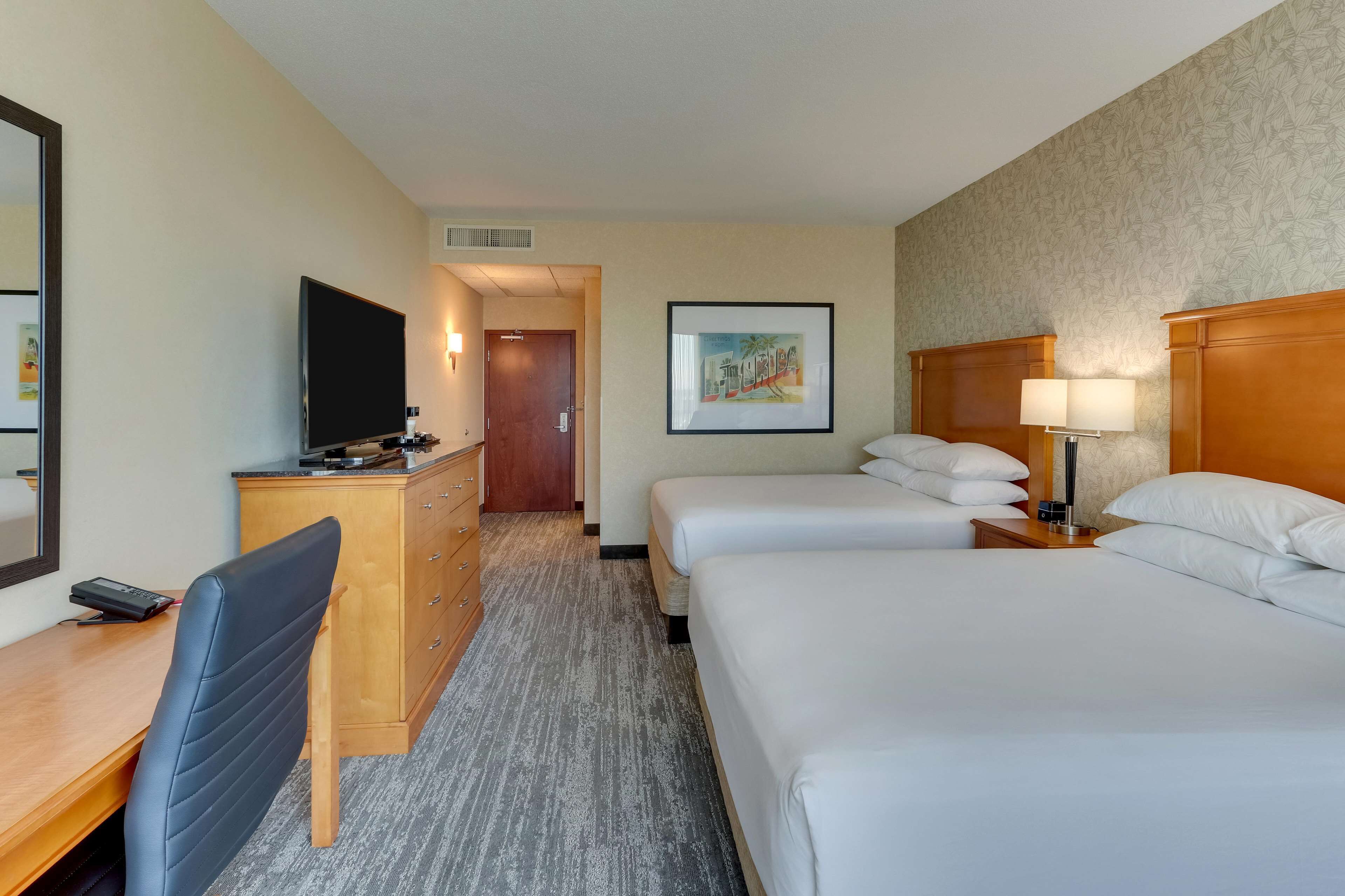 A hotel room with a bed, a desk with a chair, a television, a dresser, and a framed picture on the wall.