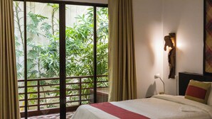 A hotel room with a bed, a nightstand, a lamp, and a view of greenery through a sliding door.