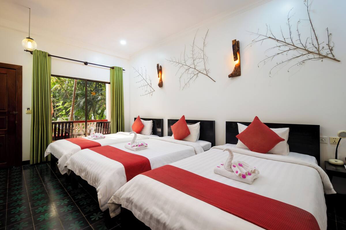 Ein Hotelzimmer mit zwei Betten, jeweils mit weißer Bettwäsche und roten Akzenten, einem Balkon mit grünen Vorhängen sowie Wanddekoration.