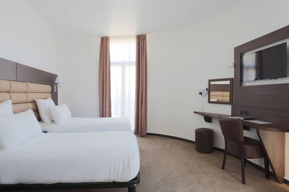 A hotel room with two beds, a desk, a chair, a TV, and a window with curtains.