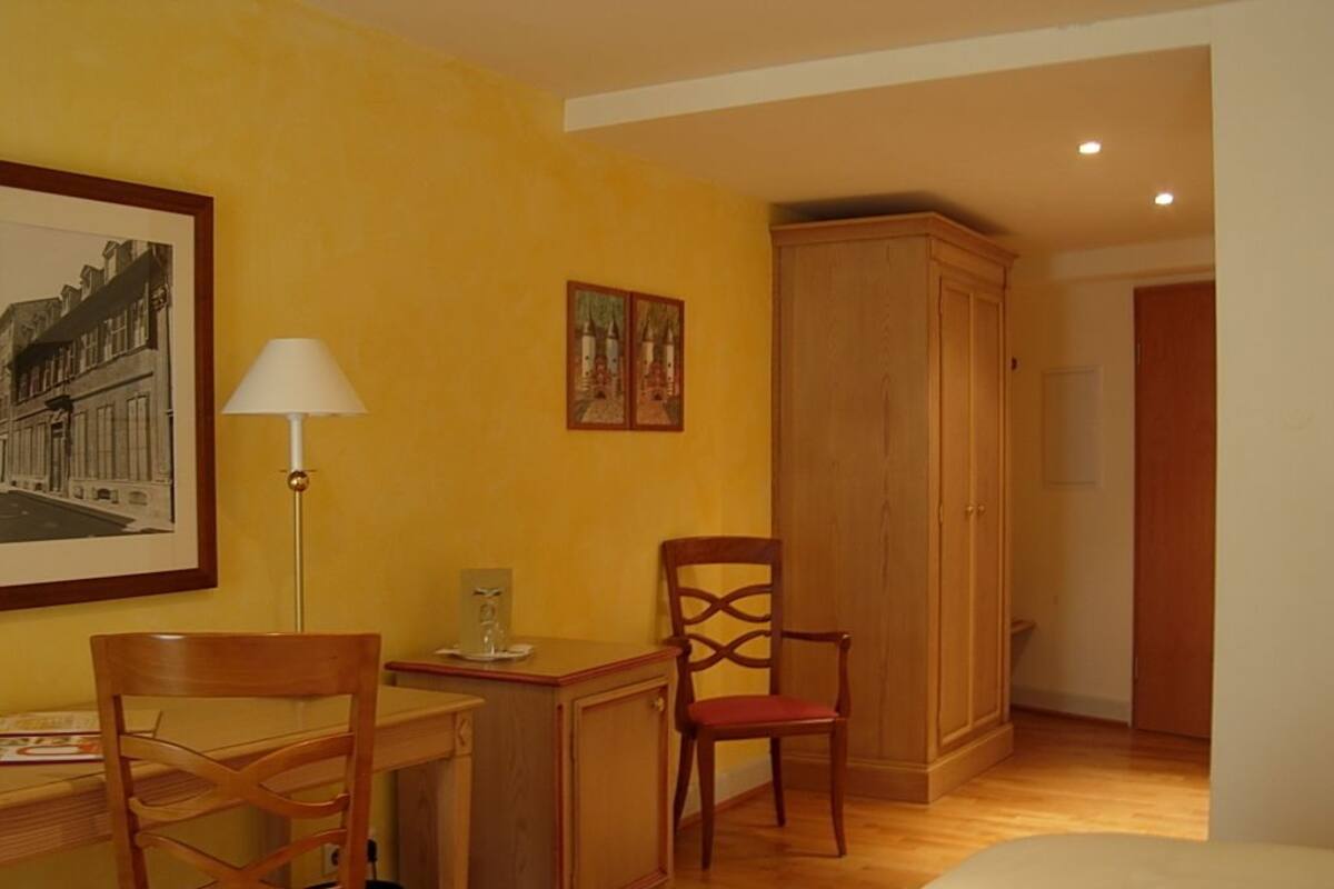 A hotel room with a bed, a desk with a lamp, a chair, a wardrobe, and a framed picture on the wall.
