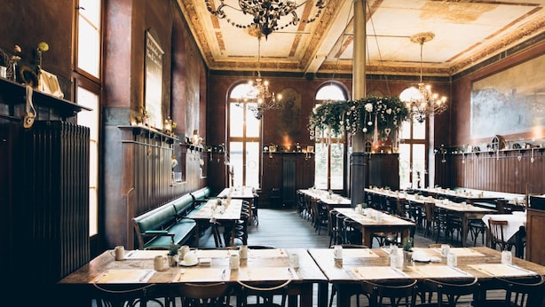 A spacious dining area with long tables and benches, large windows, and decorative chandeliers. - Kulturbrauerei Heidelberg AG (Heidelberg)