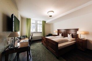 A hotel room with a large bed, two bedside tables with lamps, a desk with a chair, a television mounted on the wall, and a window with curtains. - Kulturbrauerei Heidelberg AG (Heidelberg)