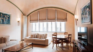 A living room with a sofa, a coffee table, a dining table with chairs, a TV, and a large window with a view. - Kulturbrauerei Heidelberg AG (Heidelberg)