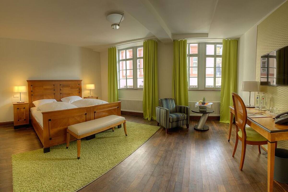 A hotel room with a wooden bed, a bench, a desk with a chair, a small table with a lamp, and a radiator.