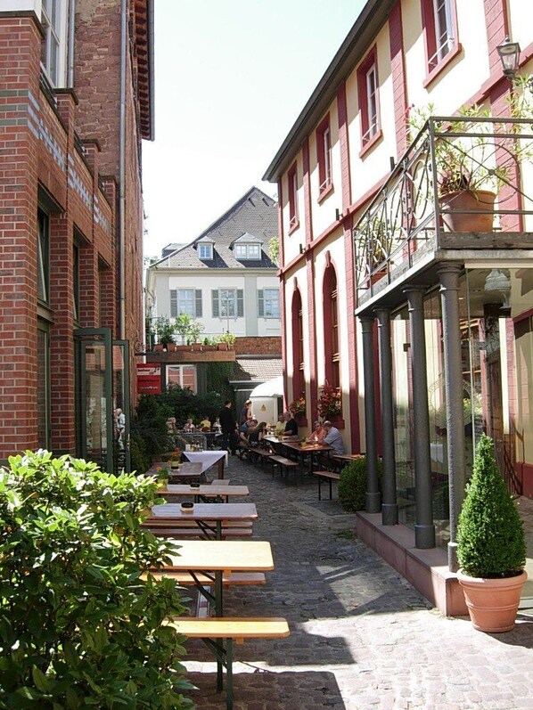 An outdoor café with tables and benches, surrounded by buildings with red brick facades and a cobblestone path.