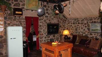 Een rustieke kamer met stenen muren, een oude koelkast, een houten tafel met fruit, een rood gordijn en een leren bank.