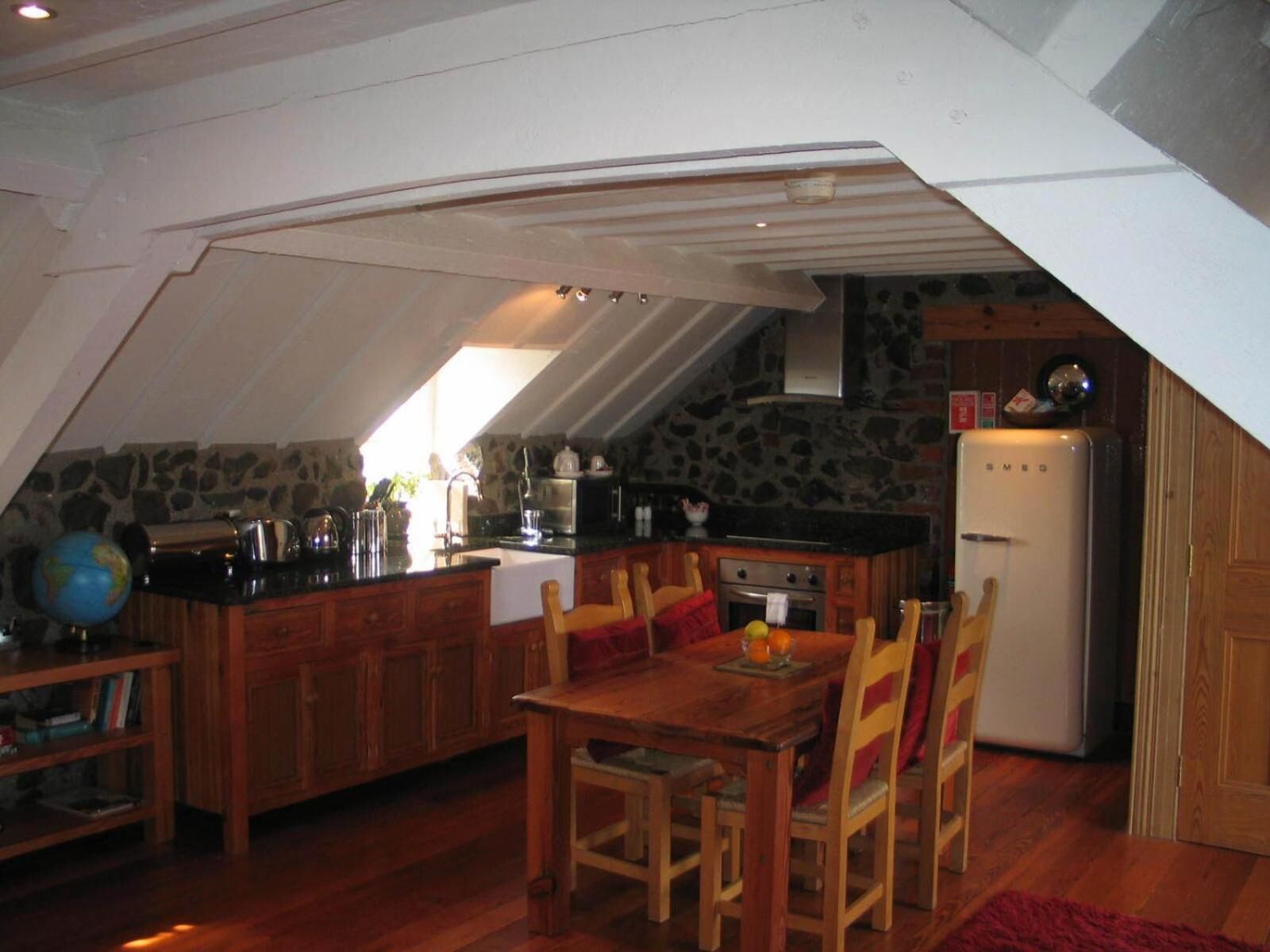 Una cocina cálida en el desván, con una mesa y sillas de madera, una pared de piedra y un refrigerador blanco.
