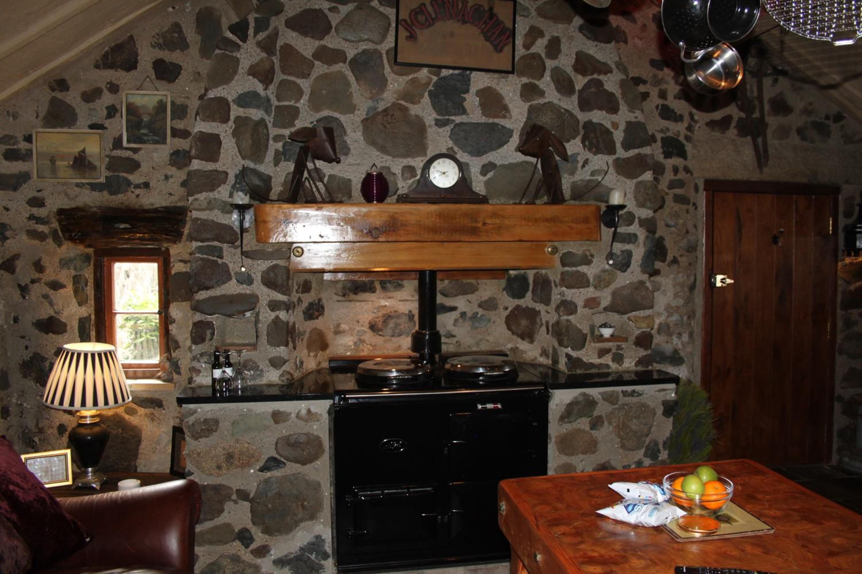 A rustic kitchen with a stone wall, wooden shelves, a stove, and a dining table with fruit.