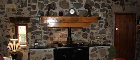 A rustic kitchen with a stone wall, wooden shelves, a stove, and a dining table with fruit.