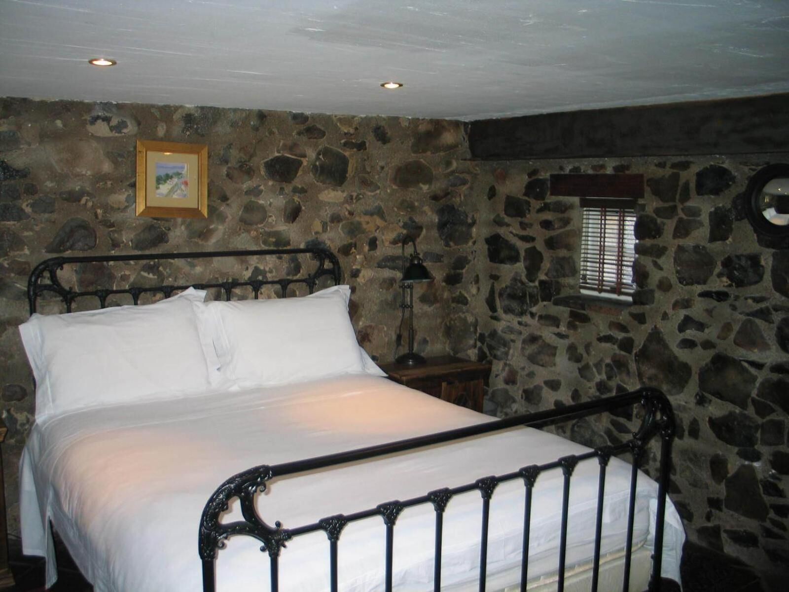 Une chambre aux murs de pierre, avec un lit en métal, une table de chevet, une lampe et une fenêtre équipée de stores.