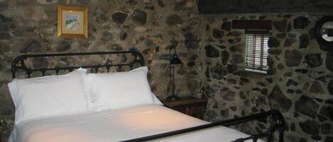 Un dormitorio con paredes de piedra, una estructura de cama de metal, una mesita de noche, una lámpara y una ventana con persianas.