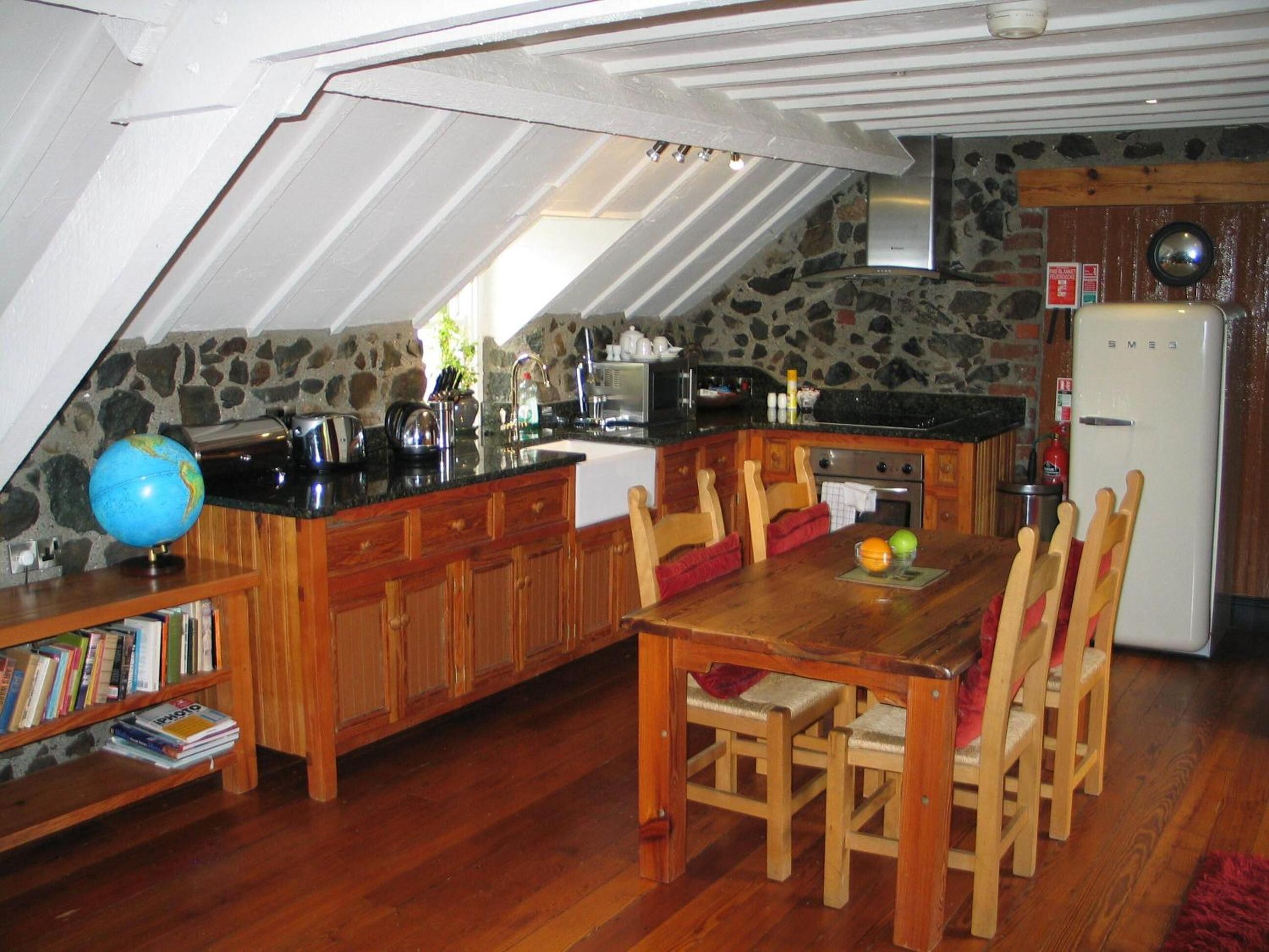 Eine gemütliche Küche mit Holzschränken, einer schwarzen Arbeitsplatte und einer Steinwand. Ein Esstisch mit Stühlen, ein Globus und Bücher auf einem Regal sind ebenfalls vorhanden.