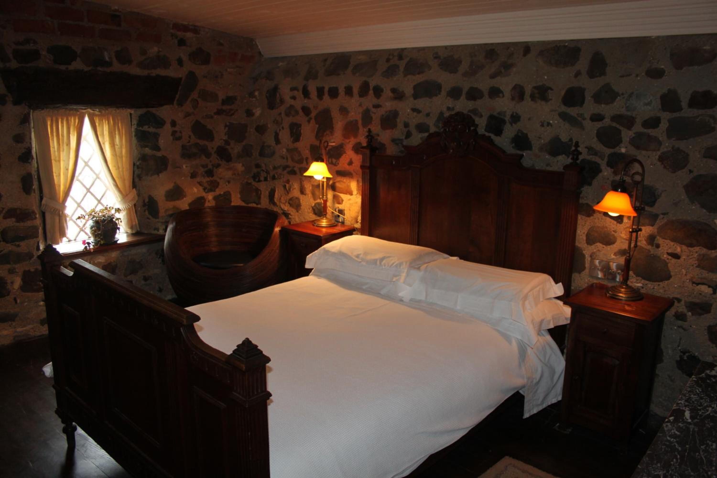 A bedroom with a stone wall, a wooden bed, two bedside tables with lamps, a chair, and a window with curtains.