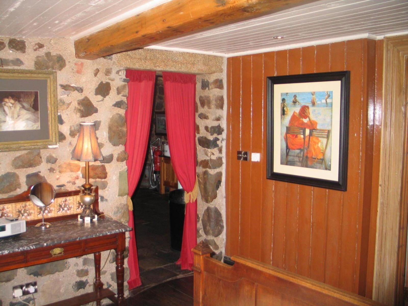 Une pièce avec un mur en pierre, une porte en bois, un tableau encadré, un bureau avec une lampe et des rideaux rouges.