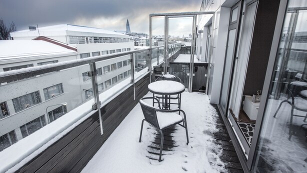 Un balcón con una mesa y silla cubiertas de nieve, con vista a un paisaje urbano con edificios y la aguja de una iglesia.