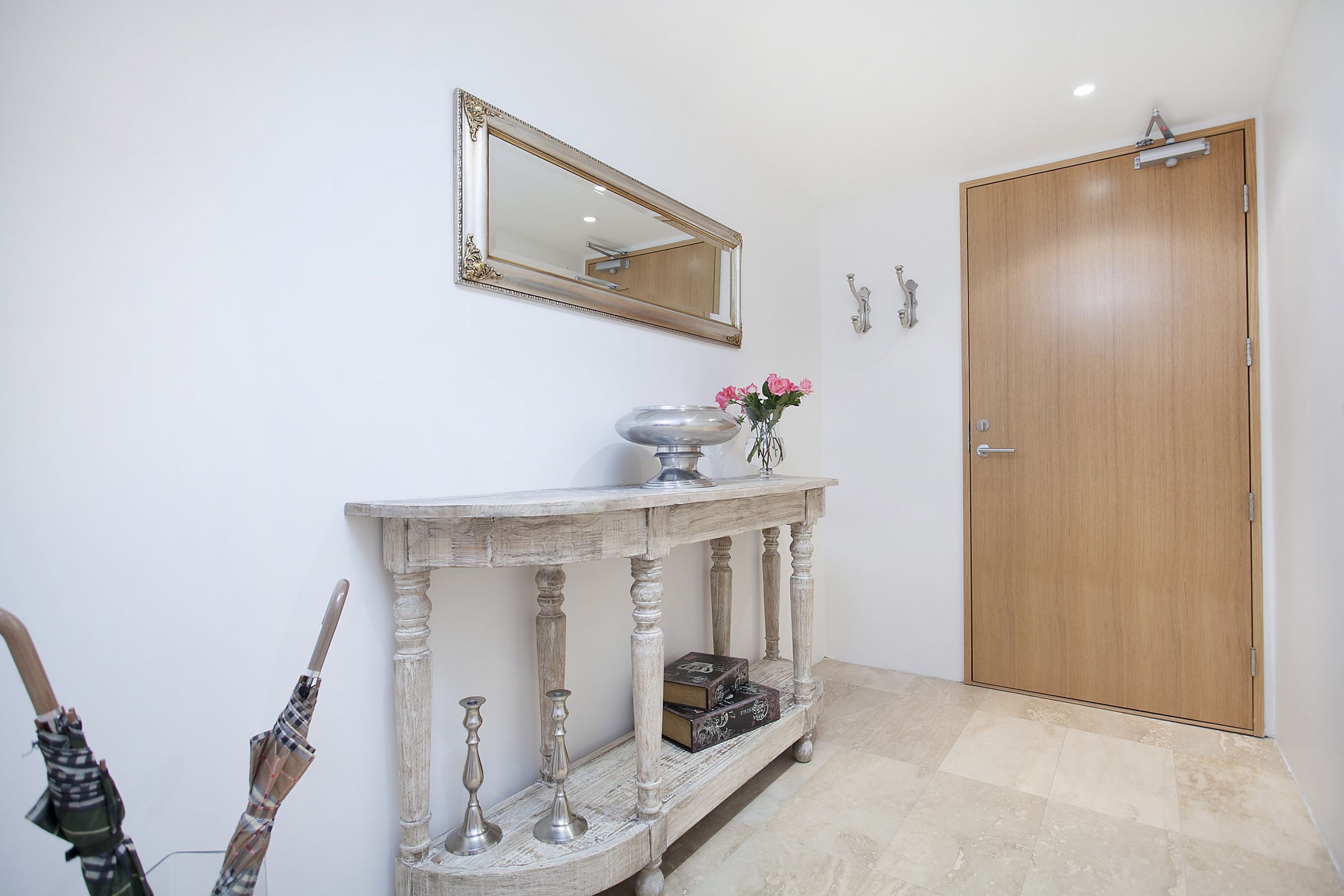 Un couloir avec une porte en bois, un miroir, une console sur laquelle est posé un vase de fleurs et un banc avec un coffre.