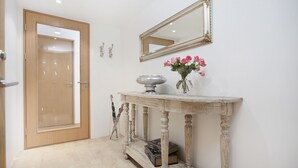 Un pasillo con una puerta de madera, una consola con espejo y un florero, y una estantería con libros.