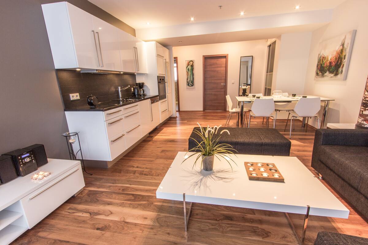 Een moderne keuken met witte kasten, een eethoek met een witte tafel en stoelen, en een woonkamer met een bank en salontafel.